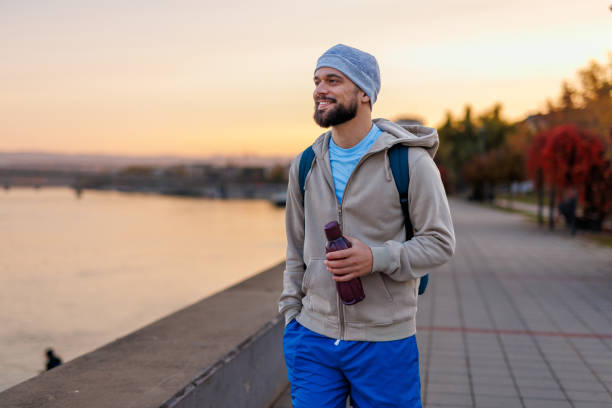 A smiling man wearing a gray hat, blue shorts, and beige hoodie holds a purple water bottle while strolling by a riverside path at sunrise, reflecting a relaxed and peaceful moment. Nature and calm surroundings enhance his morning routine.